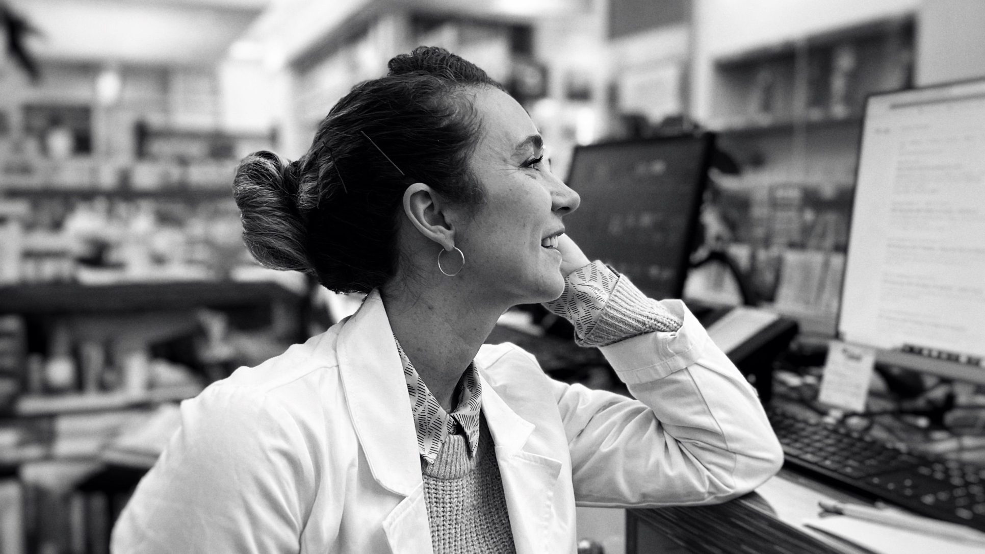 black and white photo of a female pahrmacist in a pharmacy sat in front of a computer